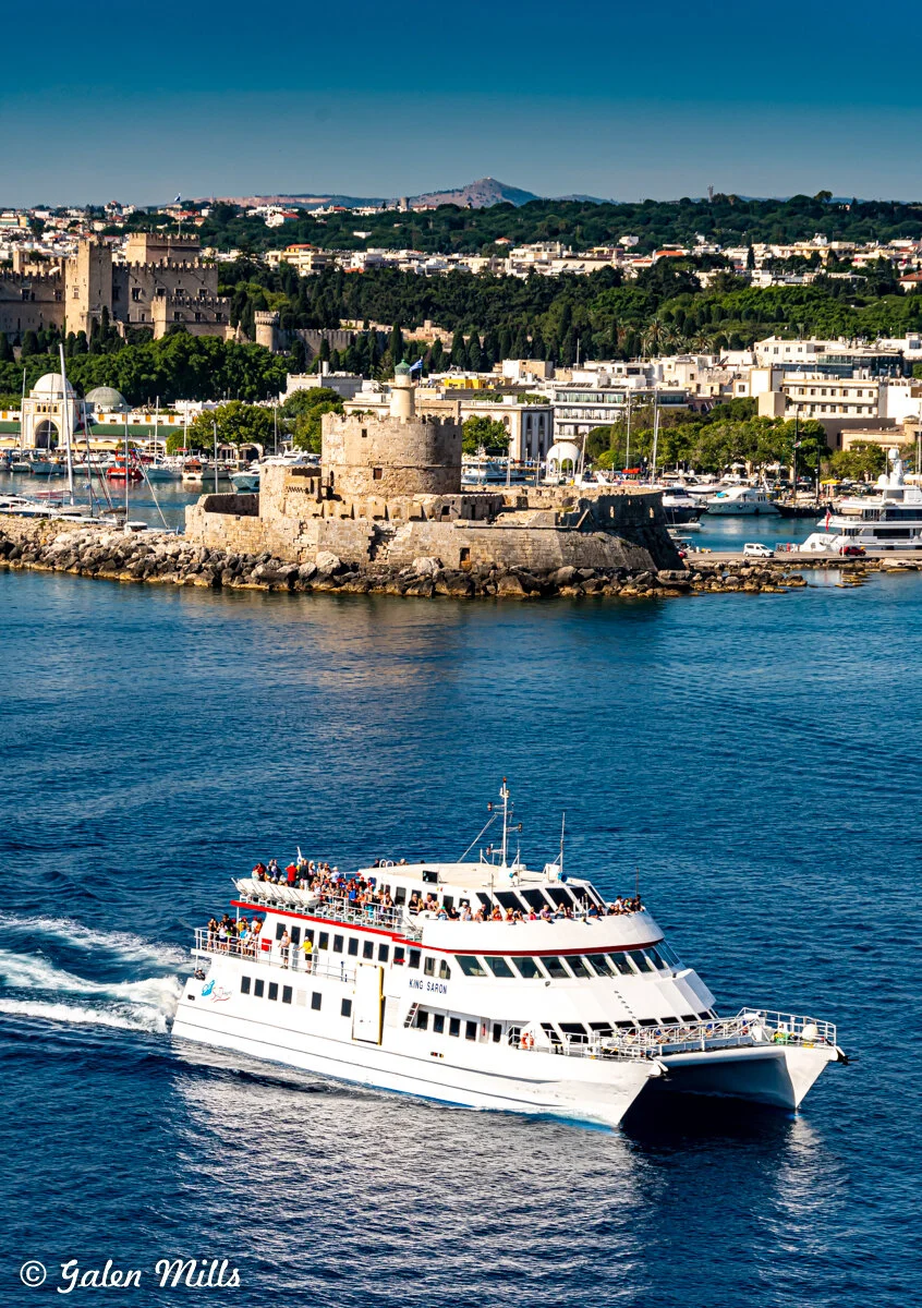 White passenger ferry sailing near medieval harbor in Rhodes, Greece with castle and town in background.
