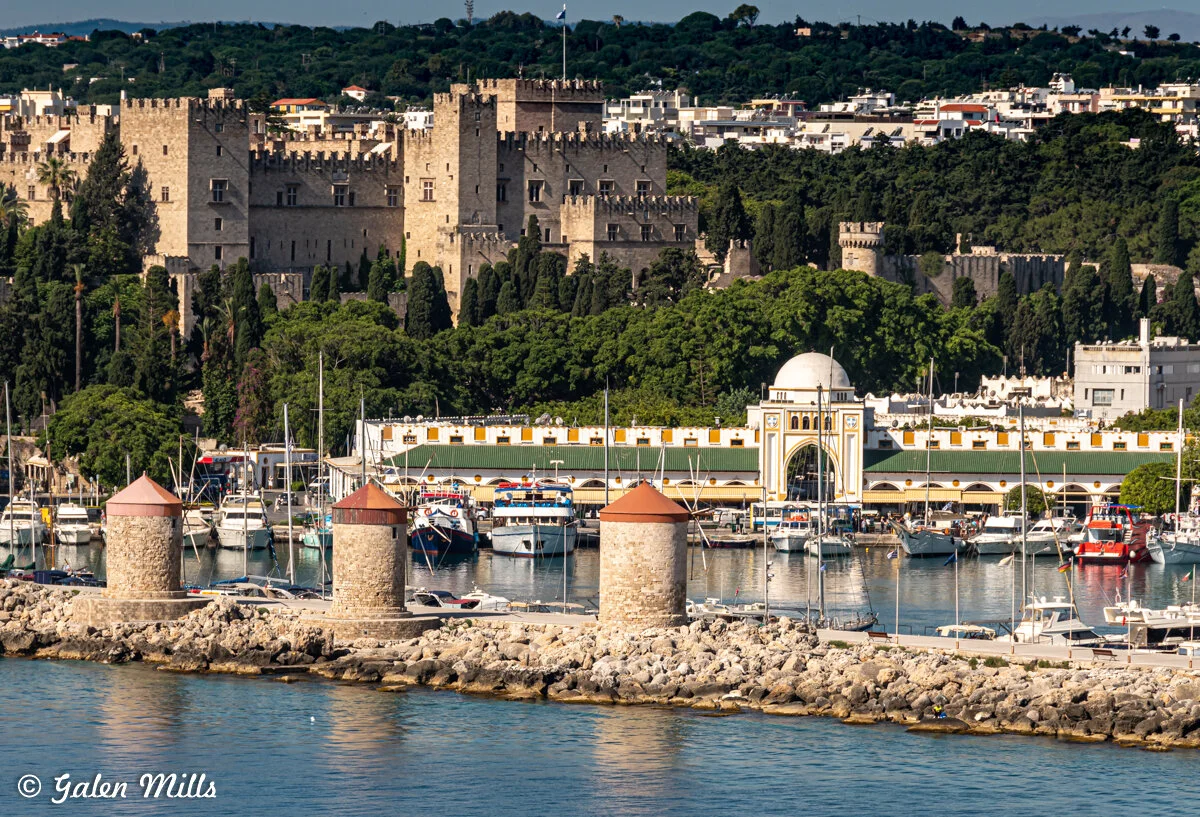 Scenic view of a historic castle with multiple towers on a lush hillside, overlooking a vibrant marina filled with boats. The foreground features a stone breakwater with several round towers and a calm blue sea.