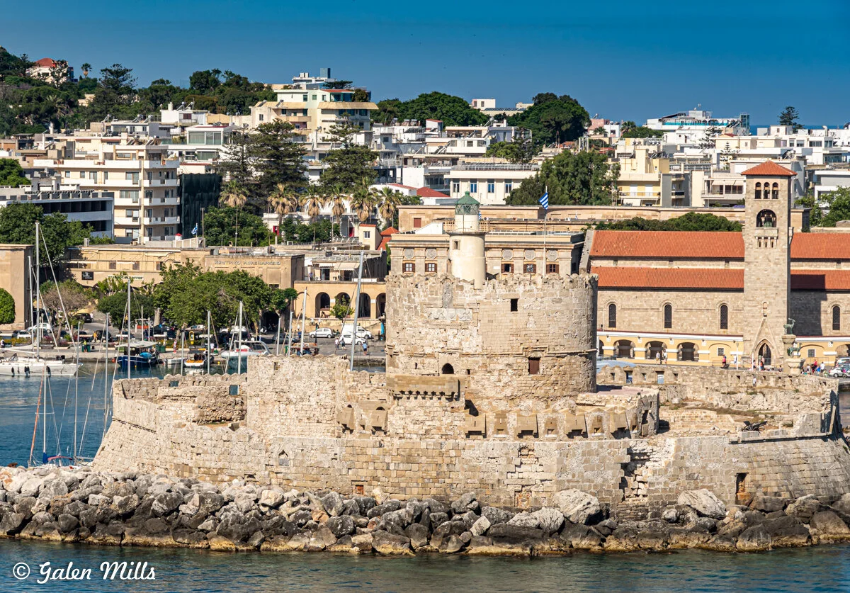 Stone fortress with lighthouse in Rhodes Harbor, Greece, surrounded by water, sailboats, and city buildings in the background.