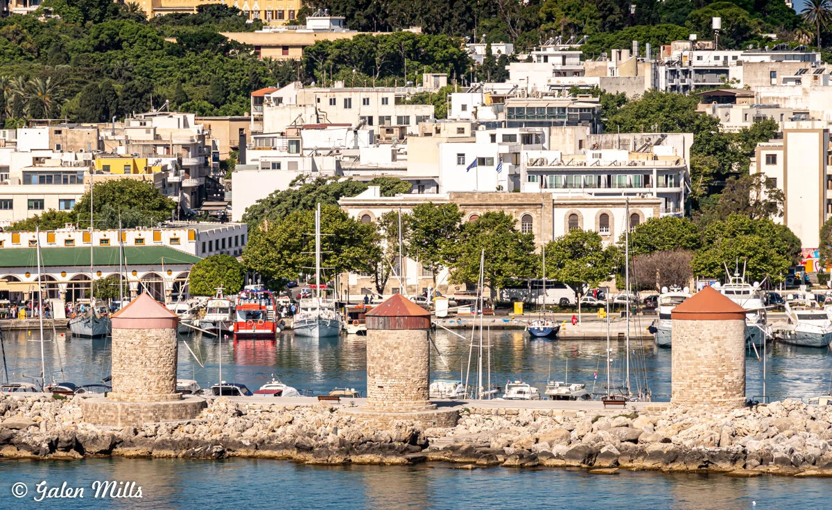 Scenic coastal view of a traditional Mediterranean town with white buildings, greenery, parked boats, and three old stone windmills by the waterfront.