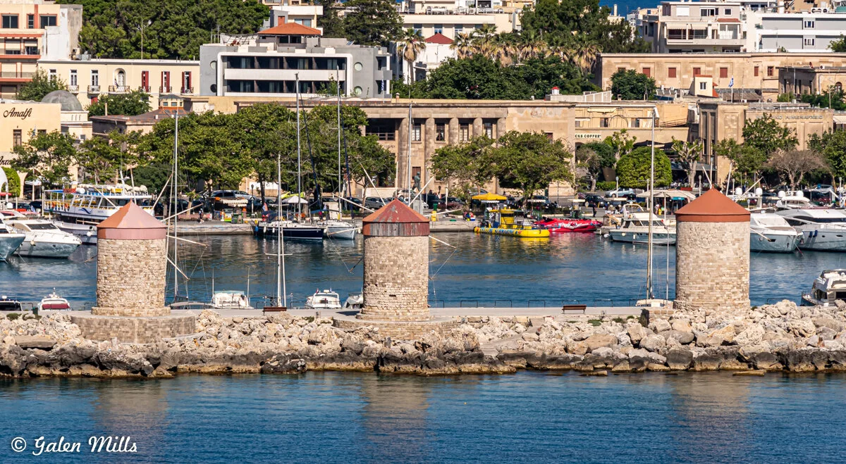 A row of three stone windmills by a harbor with boats and a cityscape in the background.