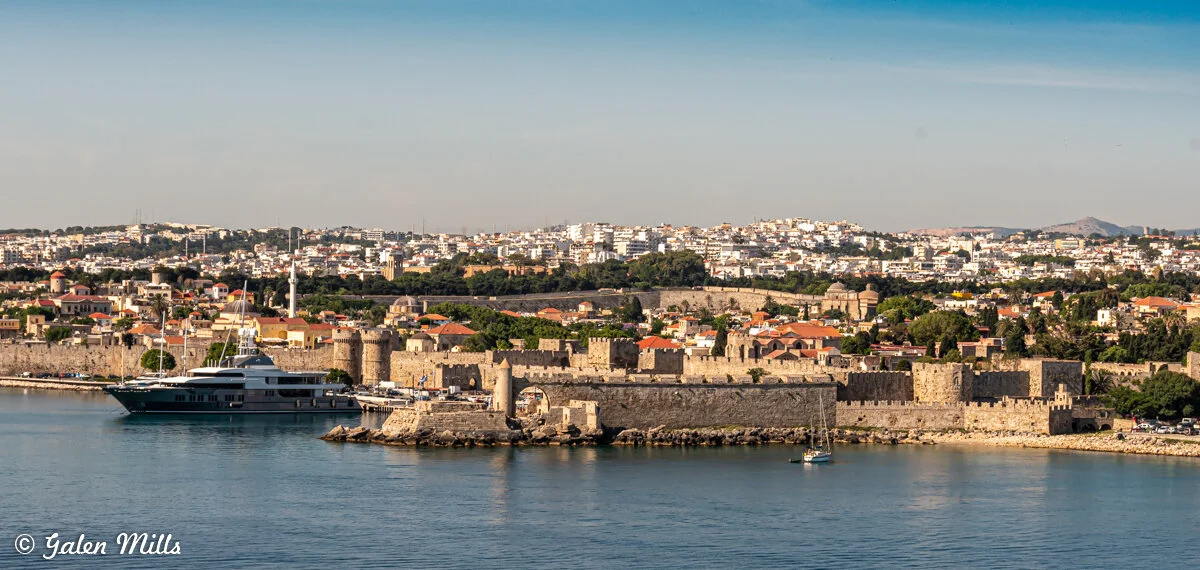 Panoramic view of a historic city with medieval walls and a harbor, featuring boats and a yacht, surrounded by modern buildings and greenery.