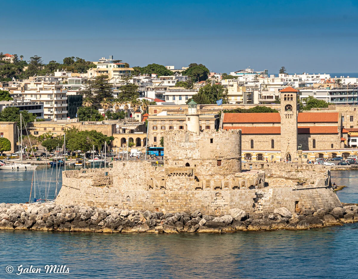 View of the Medieval Windmills and fortifications in the harbor of Rhodes, Greece, with a backdrop of modern buildings and clear blue skies.