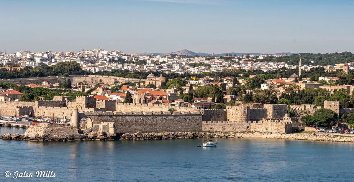 View of a historic stone fortress bordering a waterfront, with a town and hills in the background under a clear sky.