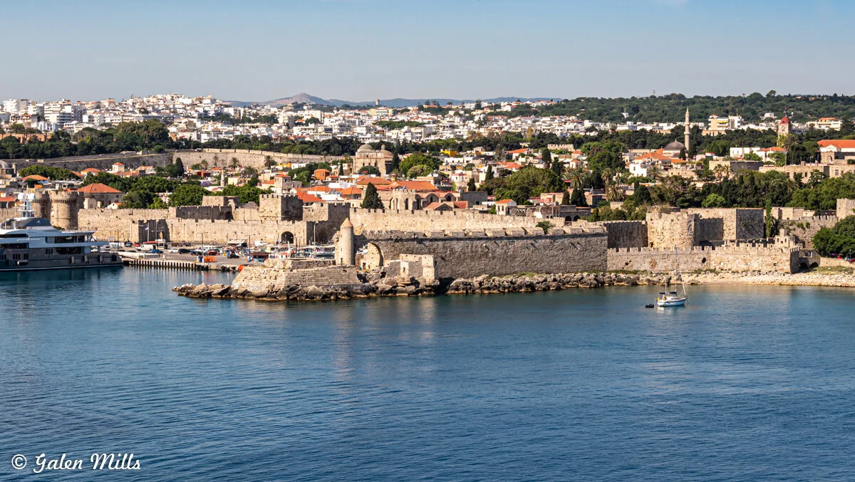 Scenic view of a historic coastal fortress in Rhodes, Greece, with boats in the harbor and a city landscape in the background.