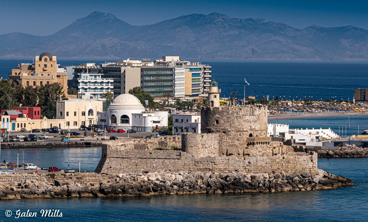 View of Mandraki Harbor in Rhodes, Greece, with historical fortification, buildings, and mountain backdrop.