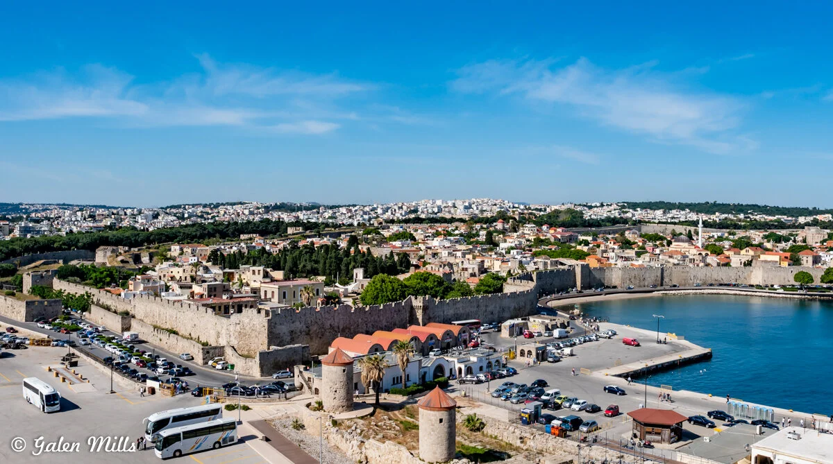 Aerial view of Rhodes Old Town, Greece, with medieval stone walls, historical buildings, and a harbor with parked vehicles and buses. The sky is clear and blue.