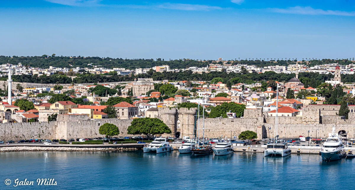 Scenic view of Rhodes, Greece featuring medieval stone walls along the coastline. Boats are moored at a marina with a cityscape of white and red-roofed buildings and green hills in the background.