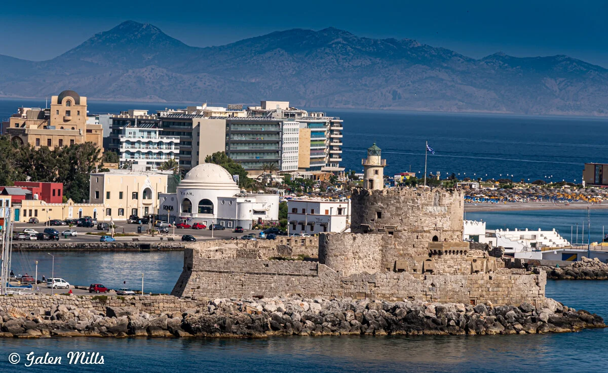 View of a coastal city with historical stone fortress, lighthouse, modern buildings, and distant mountains across the sea in daylight.