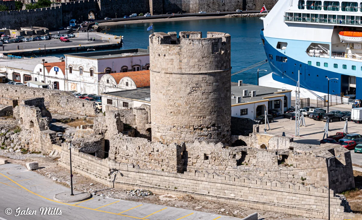 Medieval stone tower and ruins near a harbor, with a large cruise ship docked nearby. The scene includes historical architecture and modern maritime elements.