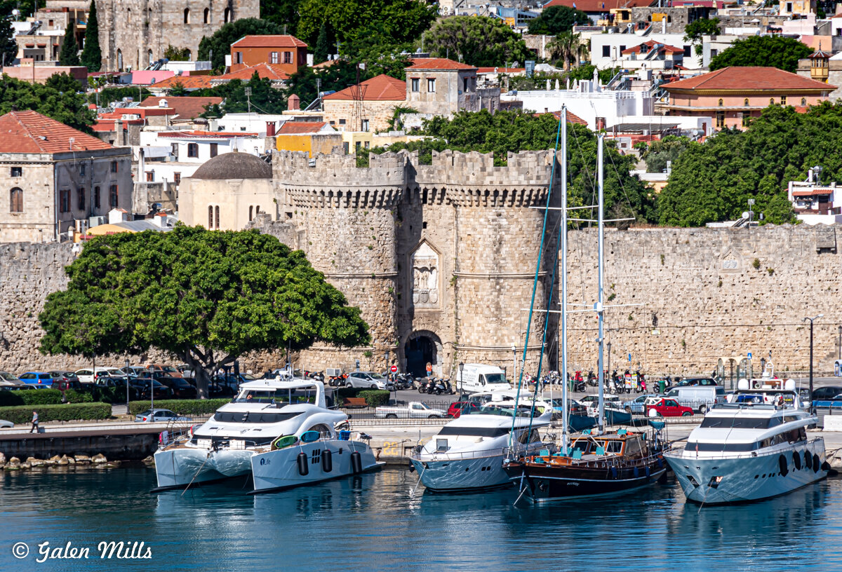 Docked yachts in a city harbor with a medieval stone fortress and residential buildings in the background.