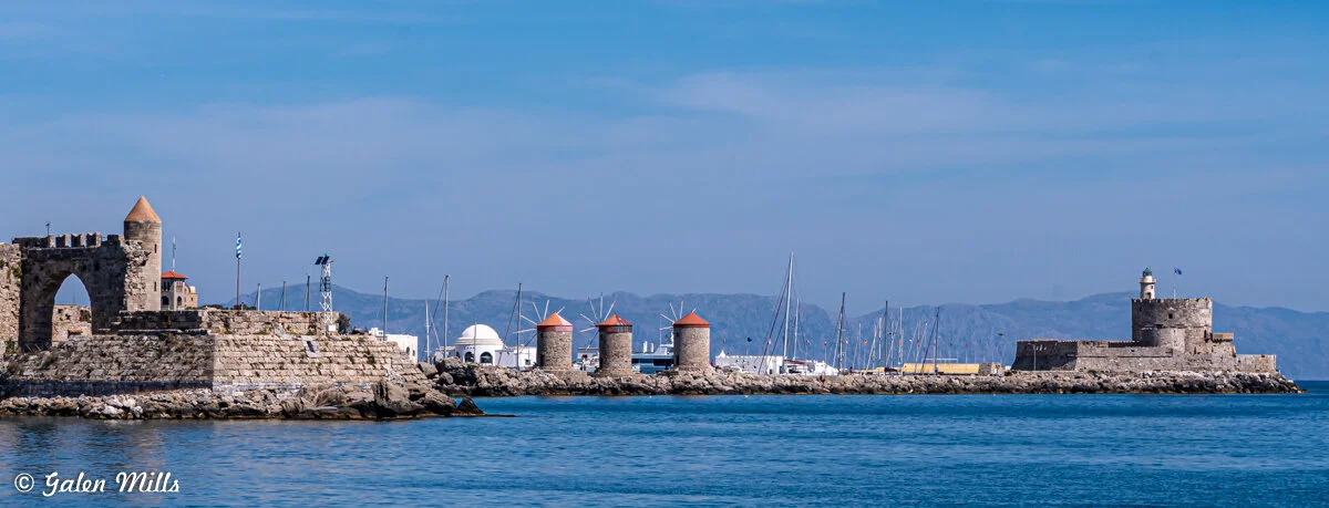 Coastal view with stone windmills on a pier, an arched fortification, and a lighthouse, against a backdrop of mountains and a clear blue sky.