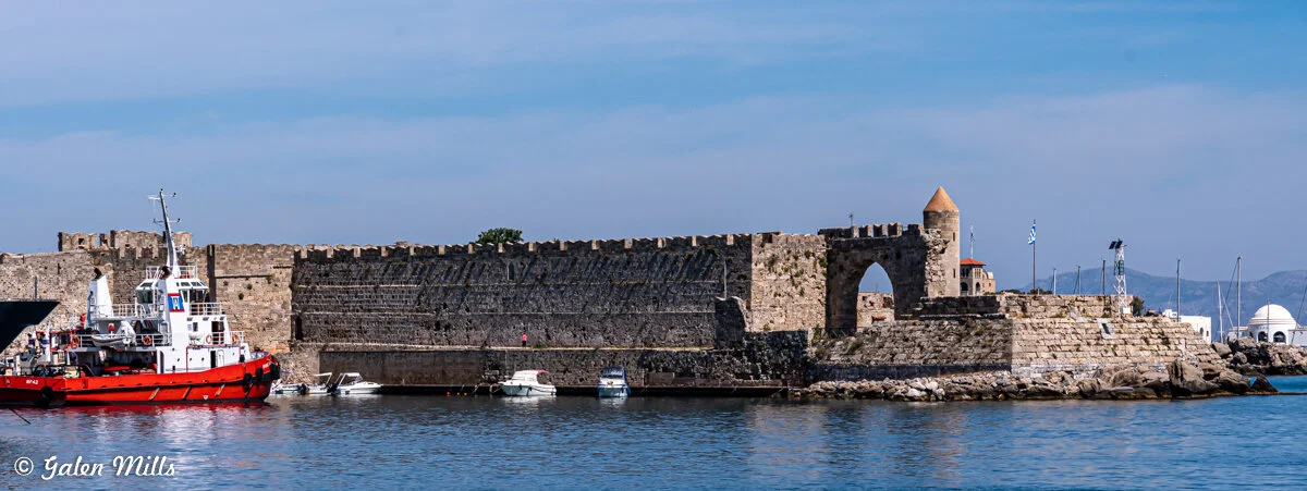 Fortified sea wall with castle-like structure and towers, red boat docked in harbor, blue sky.