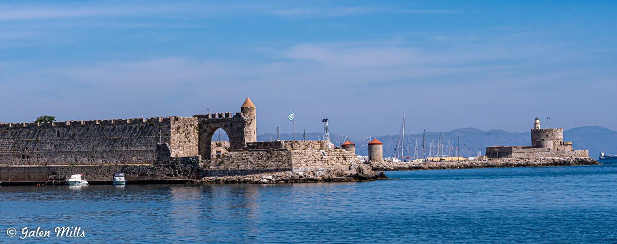 Medieval fortifications and lighthouse at the harbor entrance in Rhodes, Greece. The stone walls, towers, and Greek flag are visible by the blue sea under a clear sky.