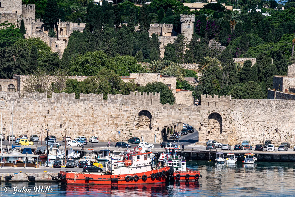 View of a historic stone wall with arched entrances, boats docked at the waterfront, and dense greenery in the background.