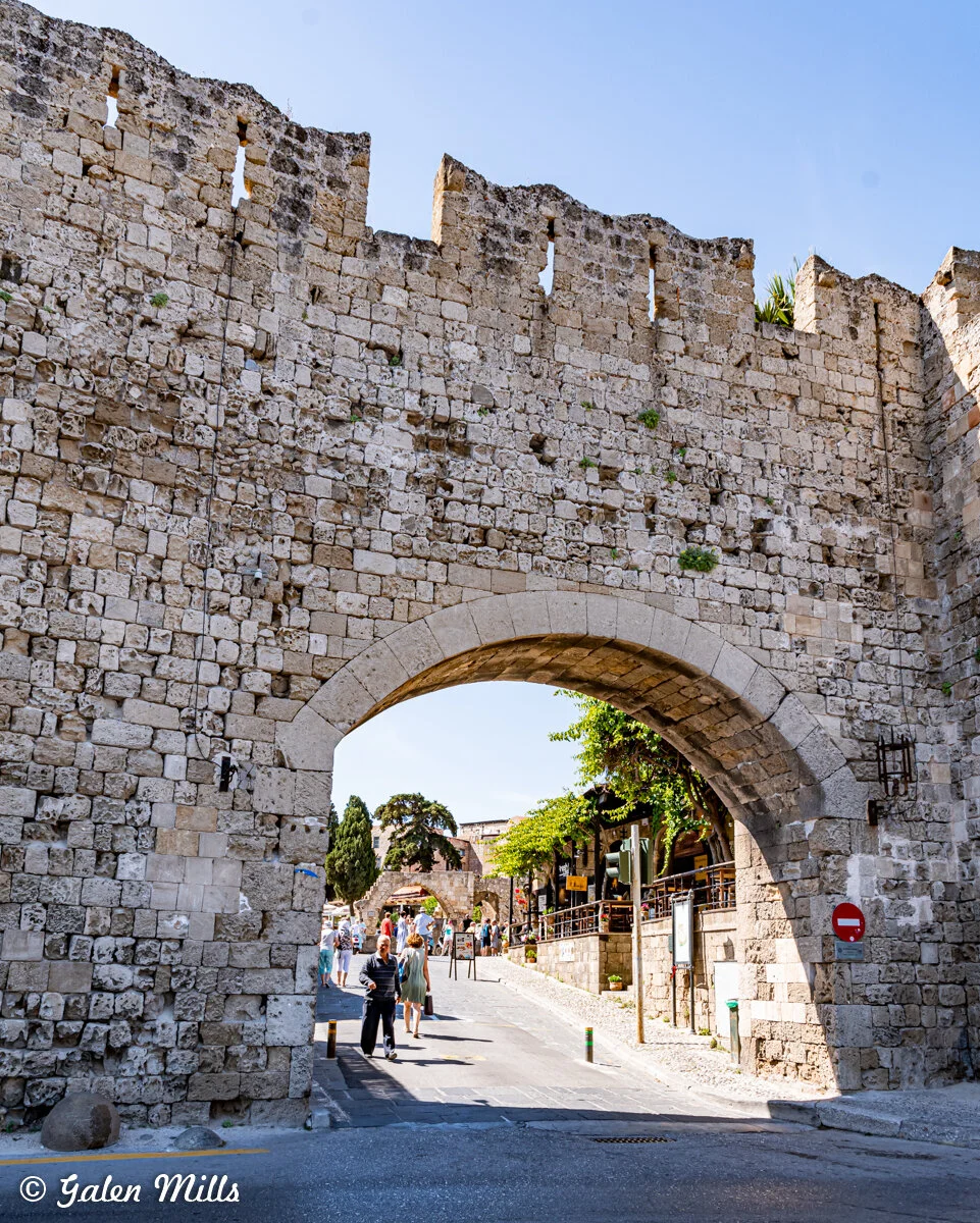 People walking through historic stone archway in medieval wall under clear sky.