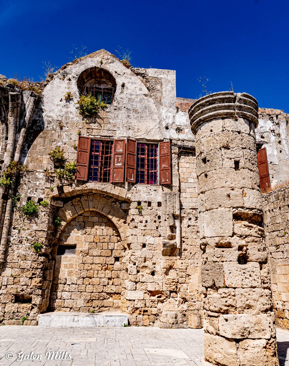 Ancient stone building façade with arched windows and red shutters, featuring a weathered stone column on a sunny day.