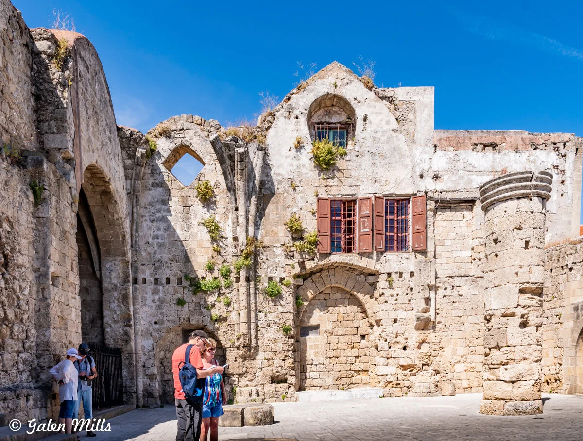 Ancient stone ruins with arches and a partially collapsed wall, featuring arched windows and exposed brickwork. A group of tourists with backpacks and hats are standing in the foreground, examining the site. Bright blue sky above.