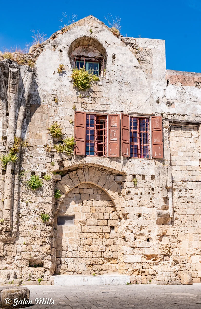 Old stone building with arched window and wooden shutters, featuring weathered stone and scattered plant growth, under a clear blue sky.