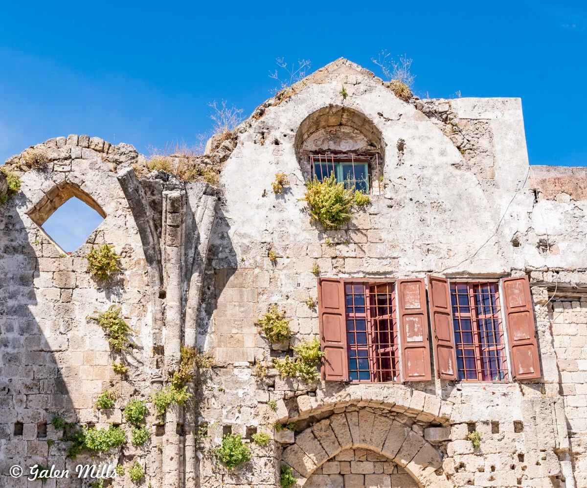 Old stone building with arched windows, partially covered in vegetation, against a blue sky backdrop.