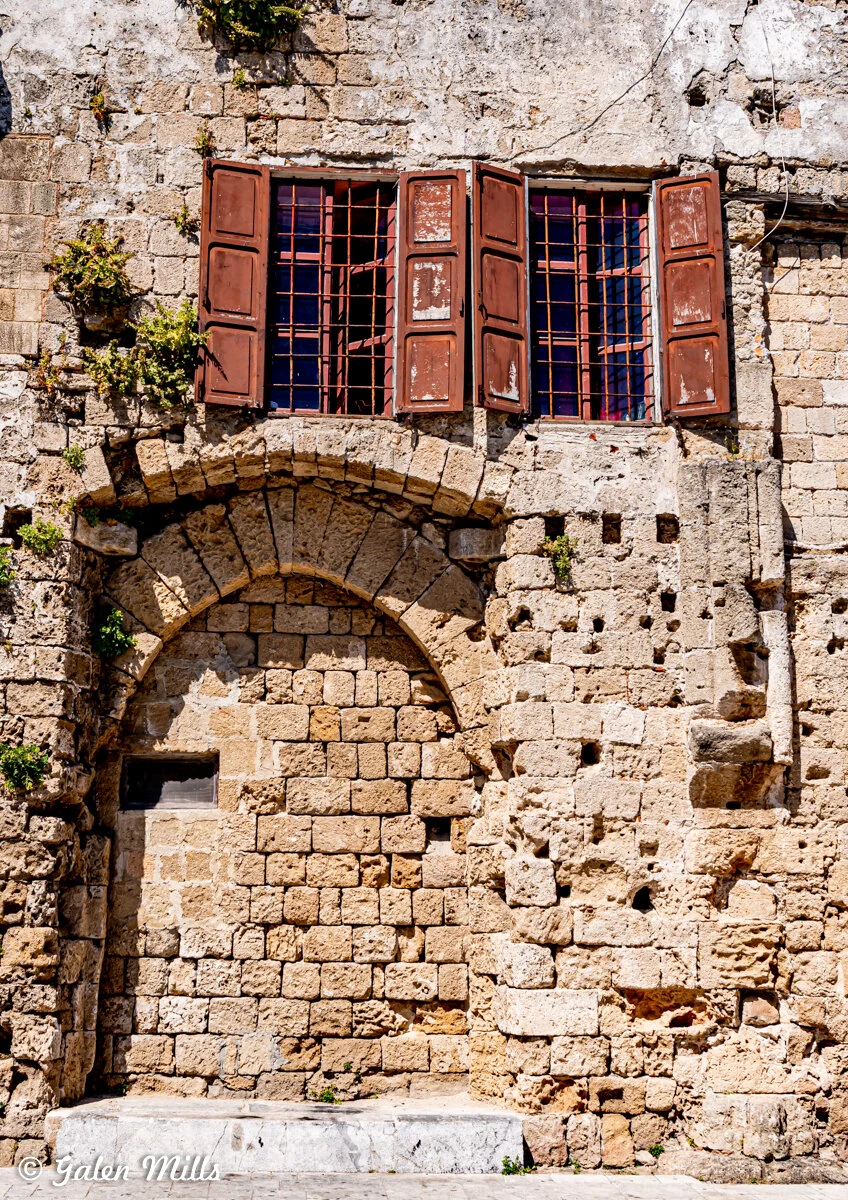 Old stone building with arched doorway and brown wooden shutters, partially open, with plants growing on the facade.