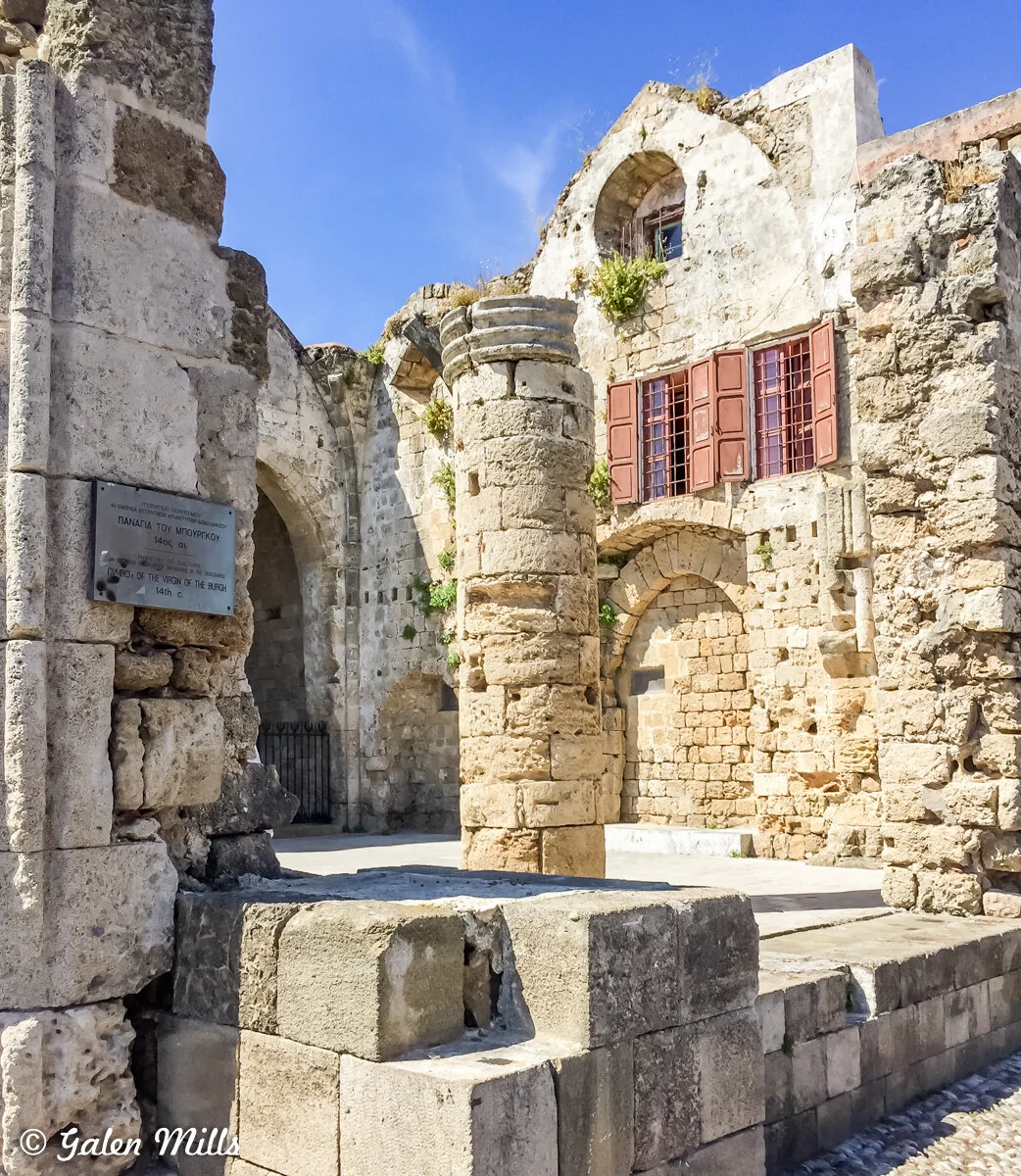 Ruins of a historical stone building with an arched entrance, ancient columns, and a weathered facade. The structure features small windows with wooden shutters and a metal plaque. Plants are growing in the crevices of the stone walls. The sky is cle