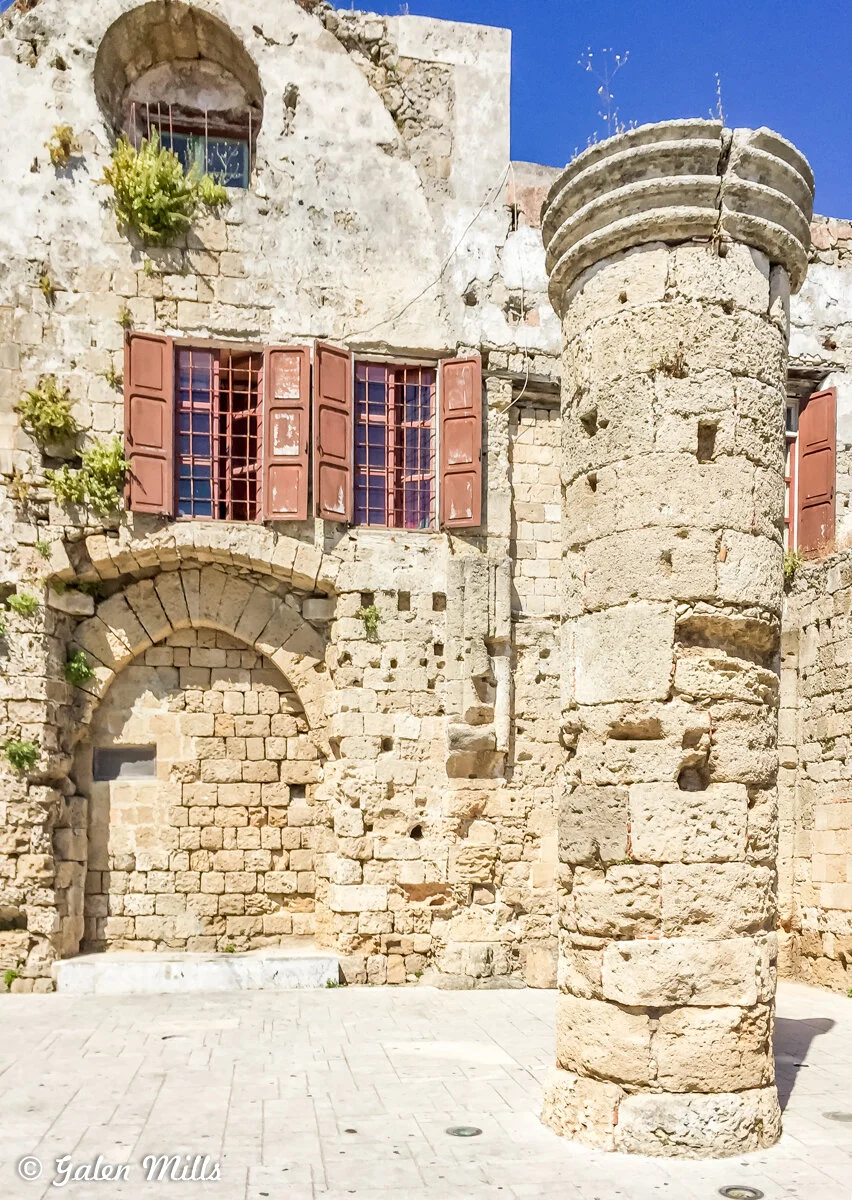 Ancient stone building with weathered columns and red shutters, surrounded by blue sky.