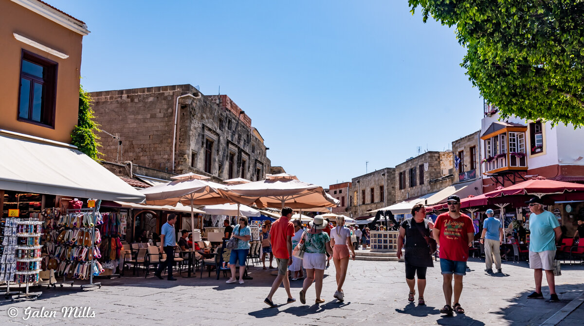 Busy street market with shops and people walking, surrounded by stone buildings and outdoor seating, under a clear blue sky.
