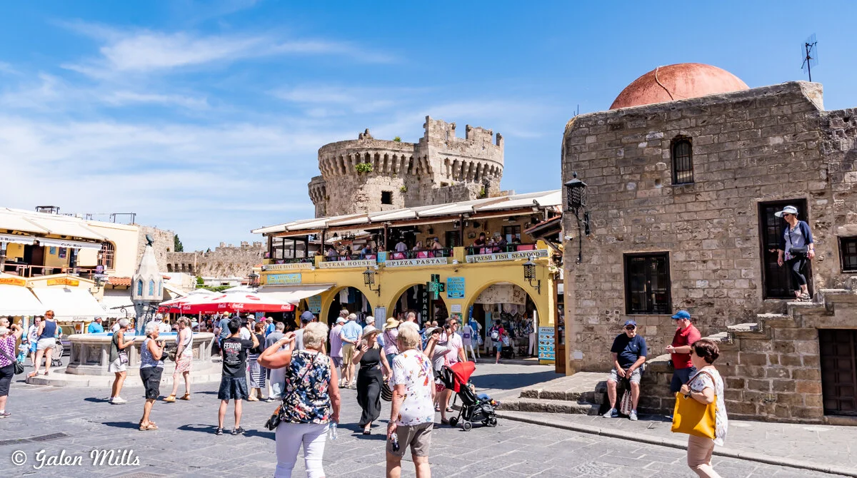Busy tourist area in Rhodes, Greece, featuring stone walls, shops, and people exploring the market under a blue sky.
