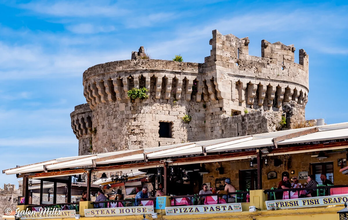 A historical stone tower with battlements is seen over a busy restaurant with outdoor seating. Signs advertise 'steak house,' 'pizza pasta,' and 'fresh fish seafood.' The sky is clear and blue.