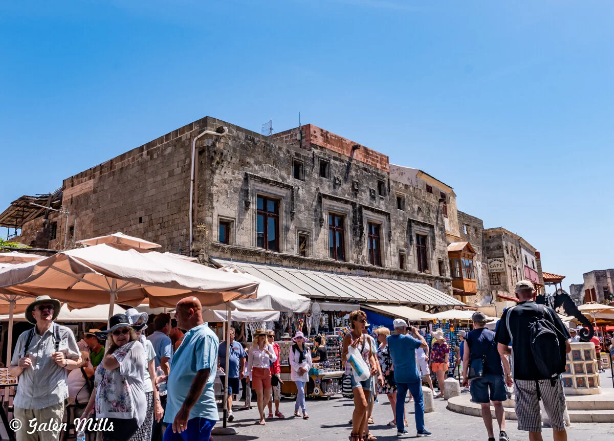 Busy outdoor market in a historic area with stone buildings and people walking under umbrellas.