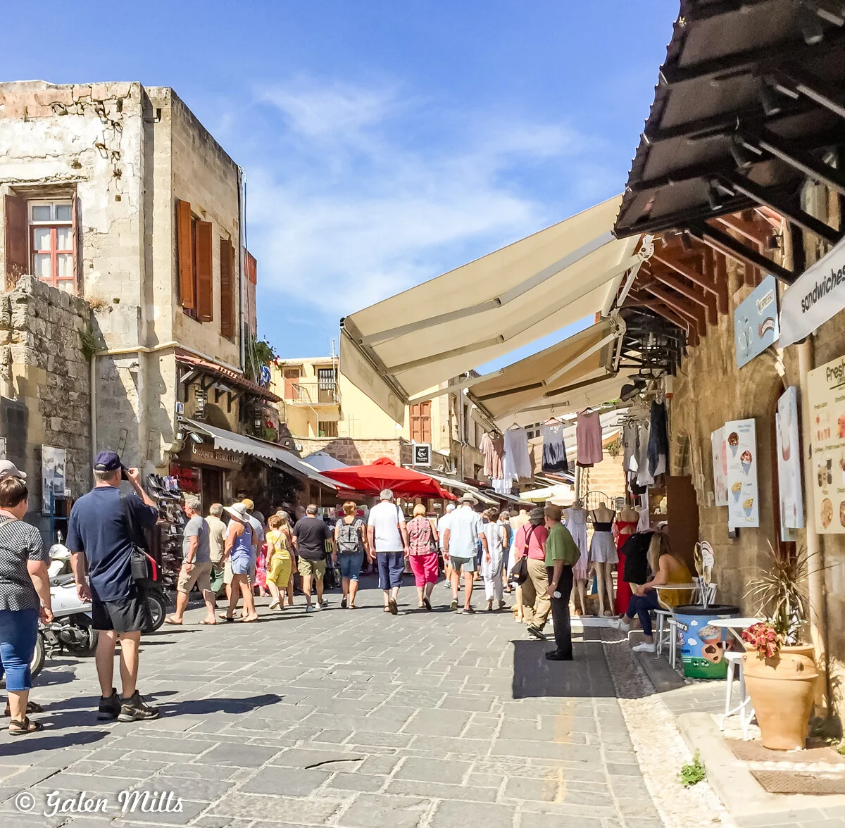 Cobbled street with people walking, lined with shops and cafes, outdoor seating, stone buildings, and overhead awnings under a clear blue sky.