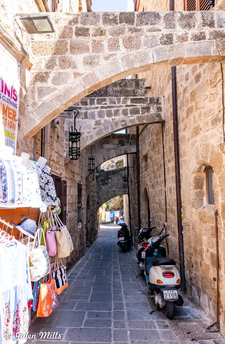 Narrow stone alleyway with arches, displaying clothing and bags on the left. Mopeds parked on the right. Bright daylight.
