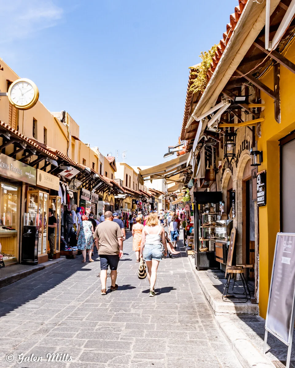 Busy pedestrian street with shops and people under a clear blue sky, featuring a prominent analog clock on the left building.