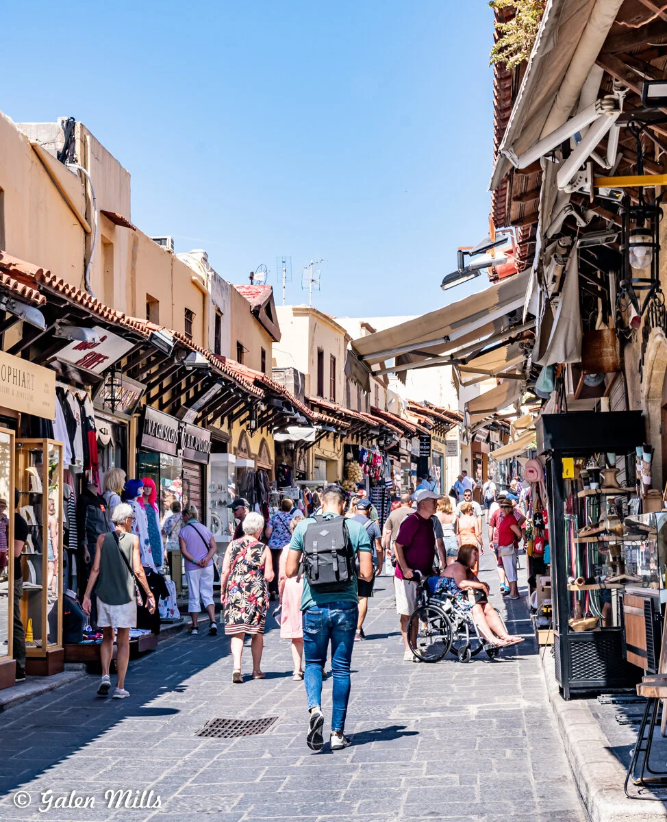 Busy outdoor market street with people shopping, wearing casual summer clothing. Shops with various goods and awnings line both sides. Blue sky in the background.