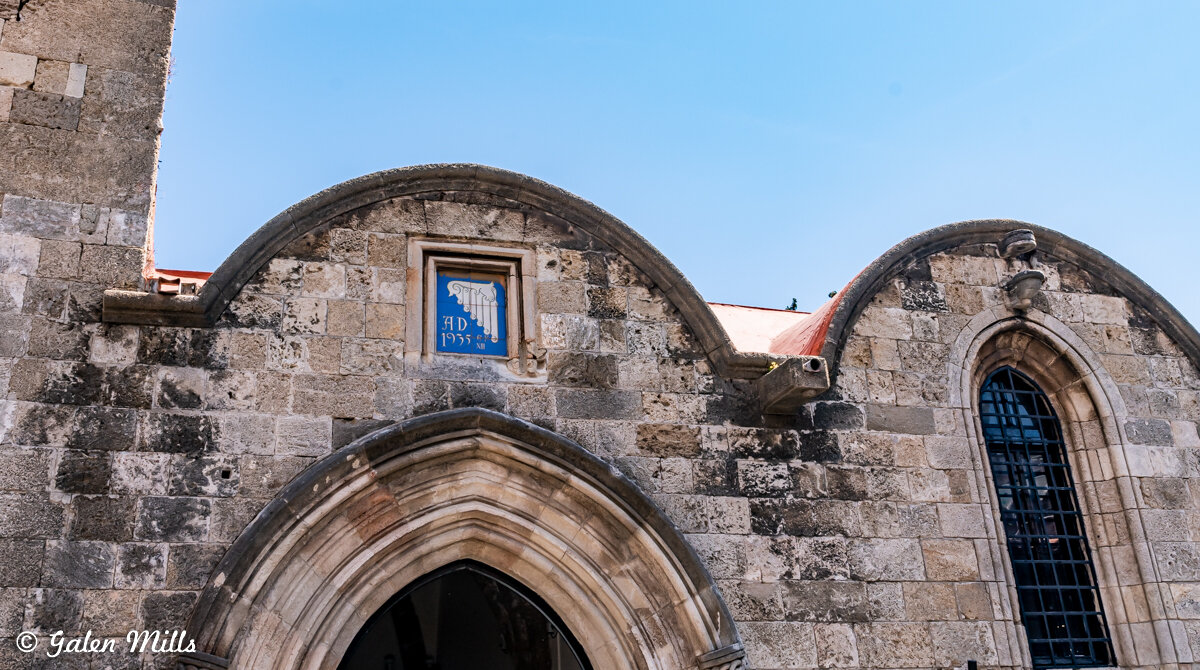 Old stone building facade with arched windows and doorway, featuring a blue plaque dated AD 1955.