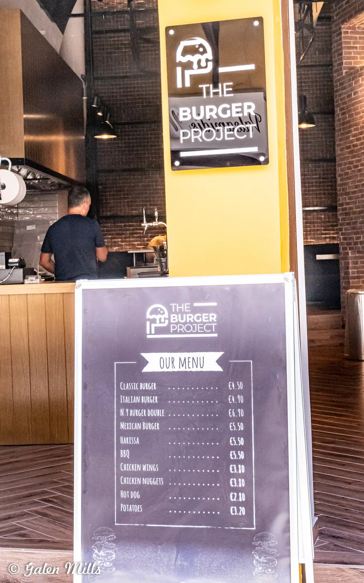 Interior of a burger restaurant with a sign reading "The Burger Project" and a menu board listing various food items and prices, including burgers, chicken wings, and hot dogs. A person in a blue shirt is visible near the counter.
