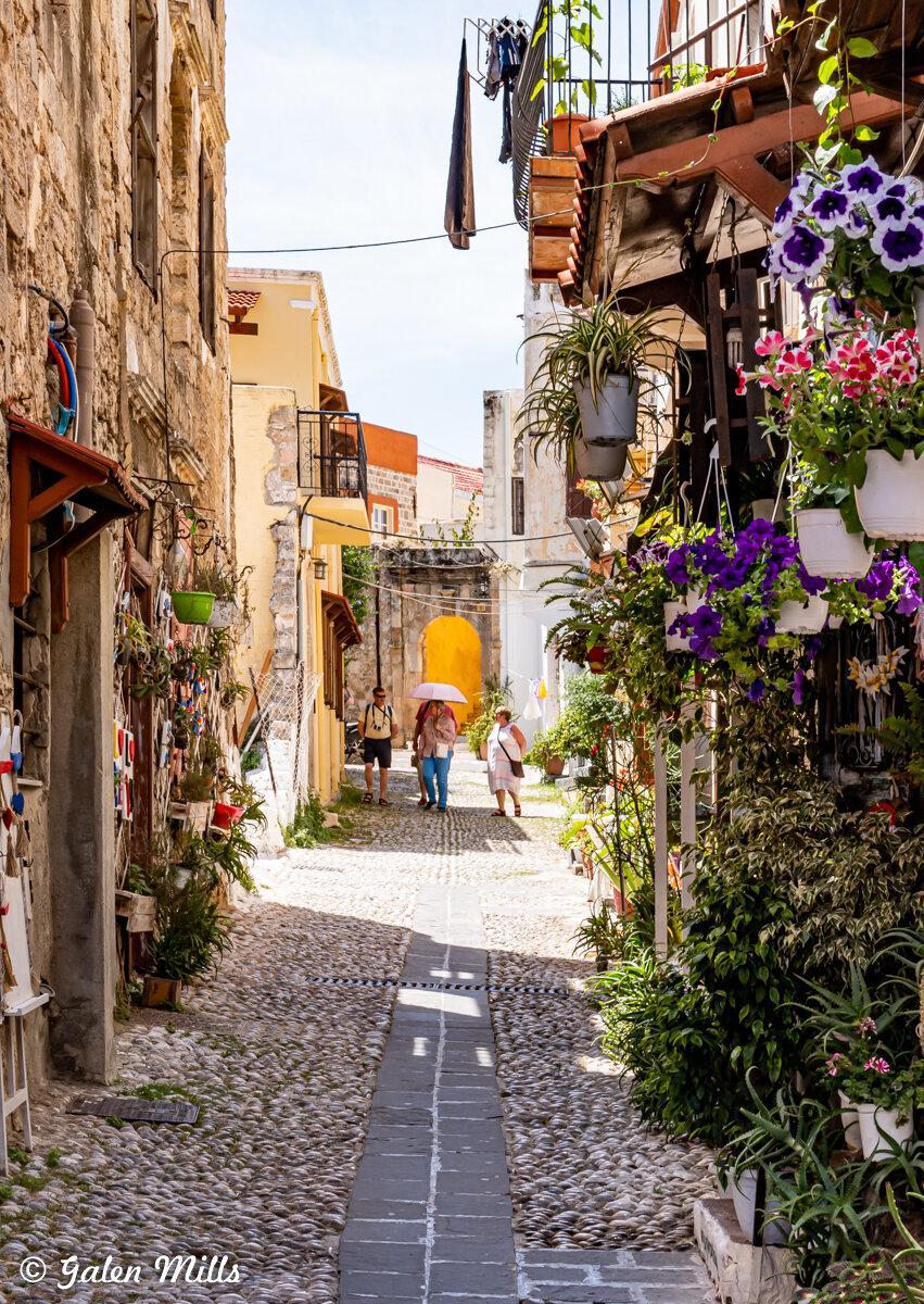 Narrow cobblestone street lined with colorful buildings and flower pots, people walking under an umbrella.