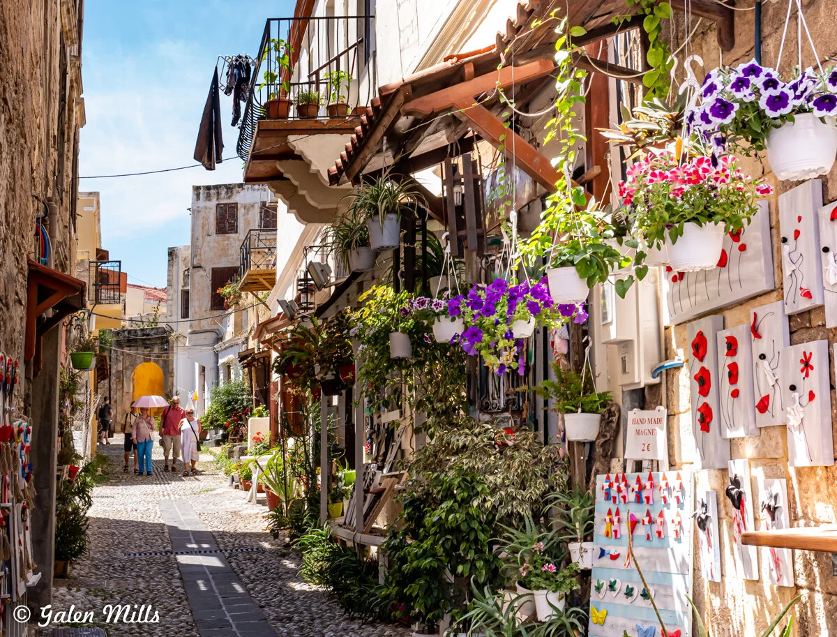 A charming narrow street in a Mediterranean town with hanging flower pots and artwork. The cobblestone path is lined with blooming plants and vibrant handmade crafts displayed along the wall. People walk in the background under a sunlit blue sky.