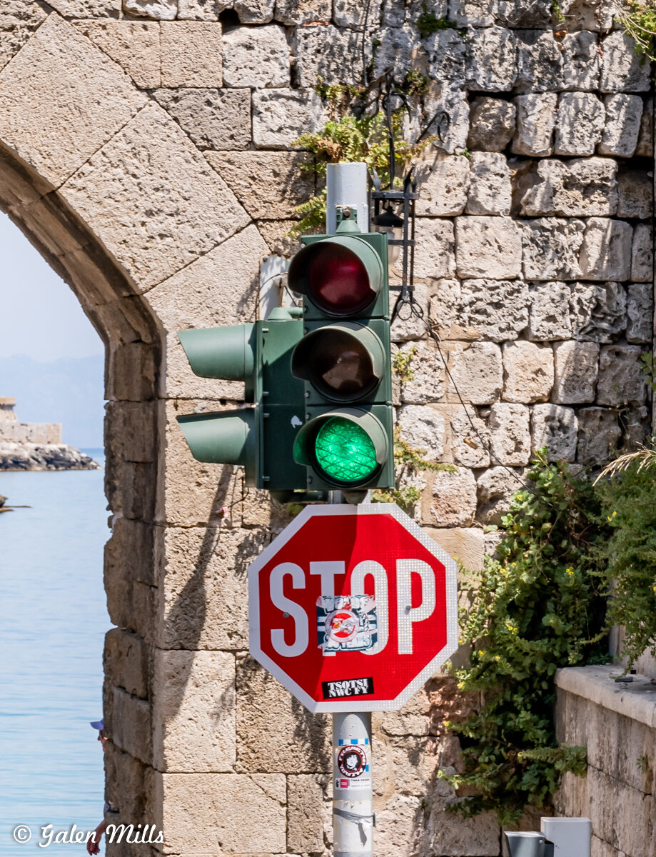 Traffic light showing green next to a stop sign with stickers, set against a stone wall background.