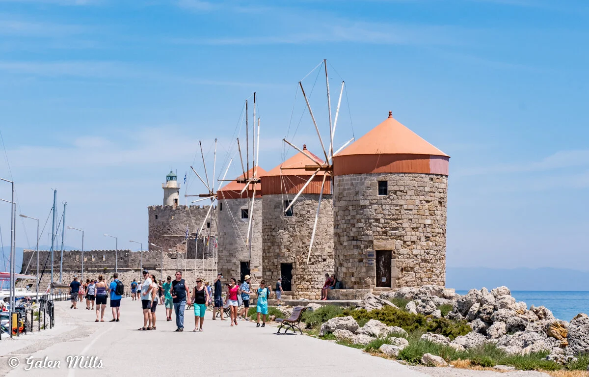 Tourists walking near medieval windmills and a lighthouse on a sunny coastal path.