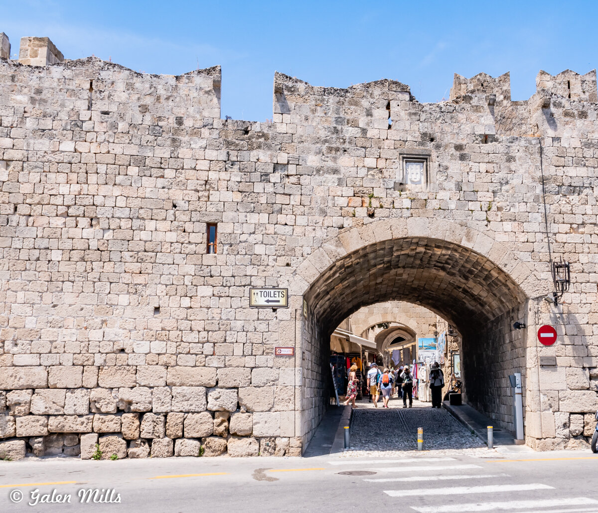 Stone archway entrance to medieval city, people walking inside, 'TOILETS' sign above, sunny day.