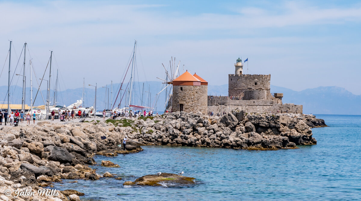 A coastal view with rocky shoreline, yachts, and a historical fortress with a lighthouse. People are walking along the pier. Bright blue sky and mountains in the background.