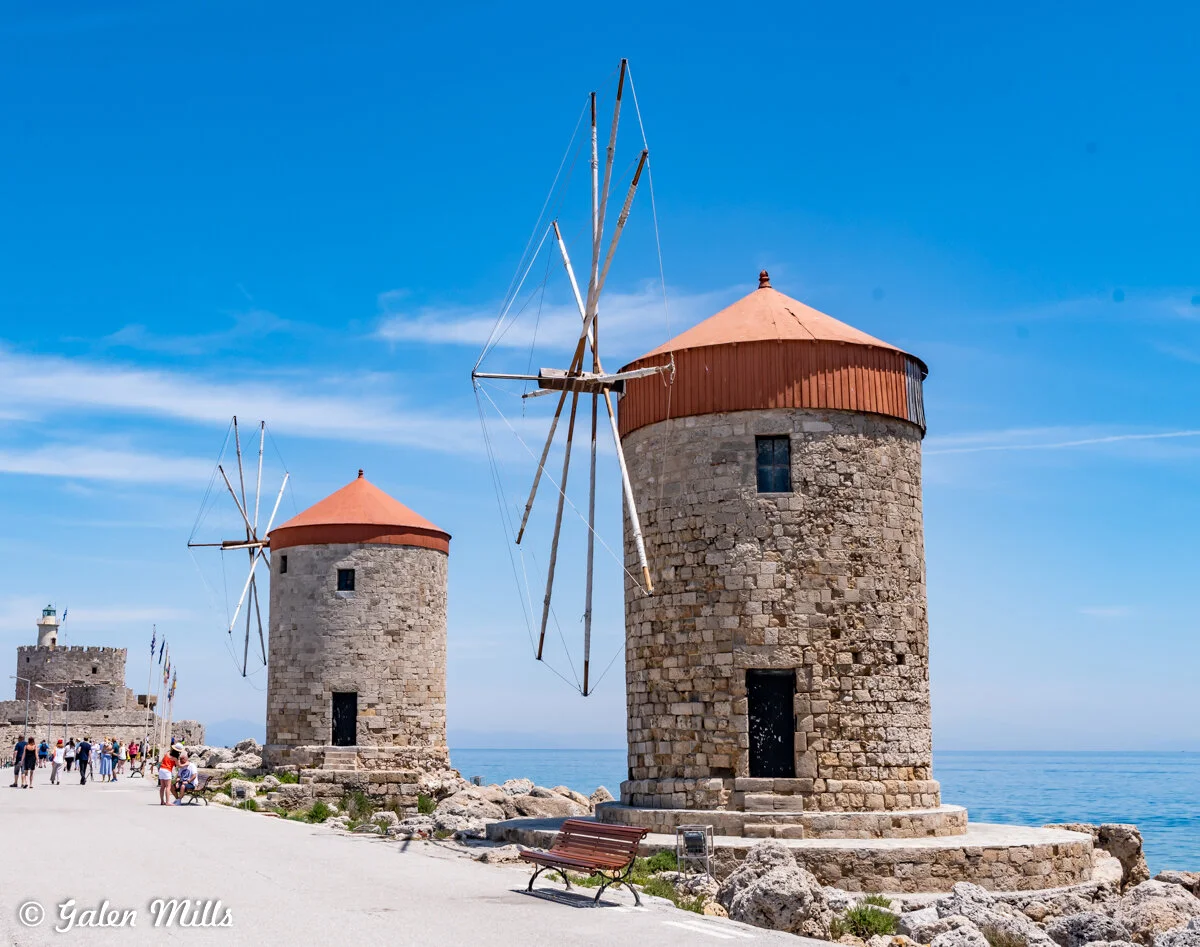 Two traditional stone windmills with red roofs near the sea, under a clear blue sky; people walking nearby.