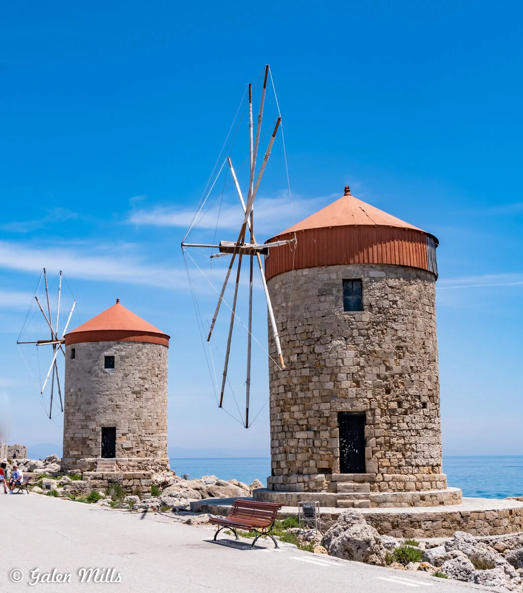 Two old stone windmills with red-topped roofs near a rocky coastline, set against a clear blue sky.