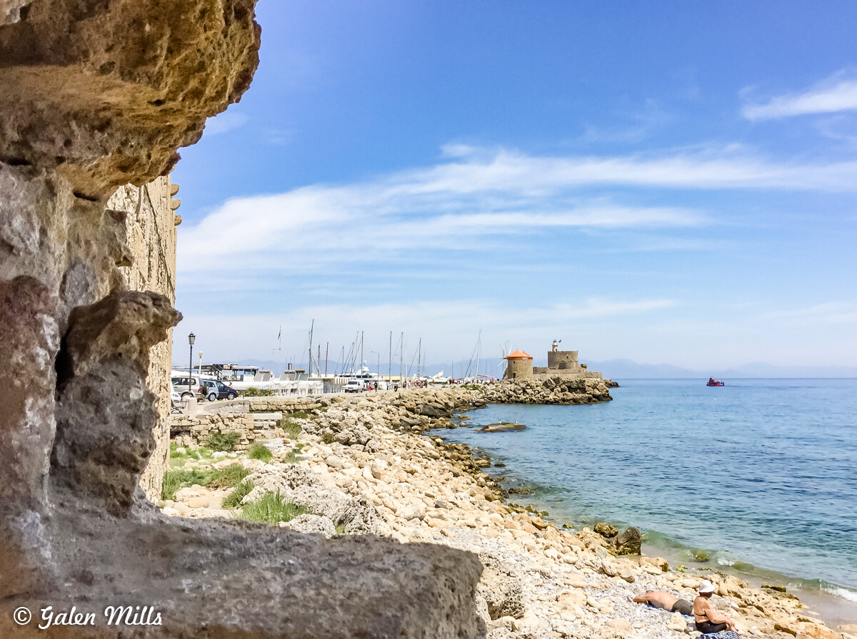 Stone wall with coastal view of beach, rocky shoreline, sea, boats, and ancient castle-like structure under a blue sky.