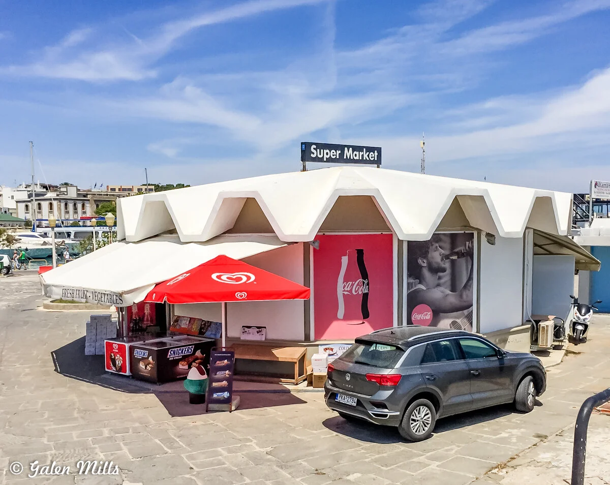 Exterior of a small supermarket with a modern white facade and zigzag roof design. A red umbrella is in front, with a sign offering fresh fruit and vegetables. Prominent advertisements feature Coca-Cola imagery, alongside stands displaying Snickers a