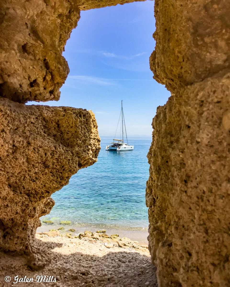 View of a sailboat on the ocean through a rock formation opening, with sandy beach and clear blue sky.