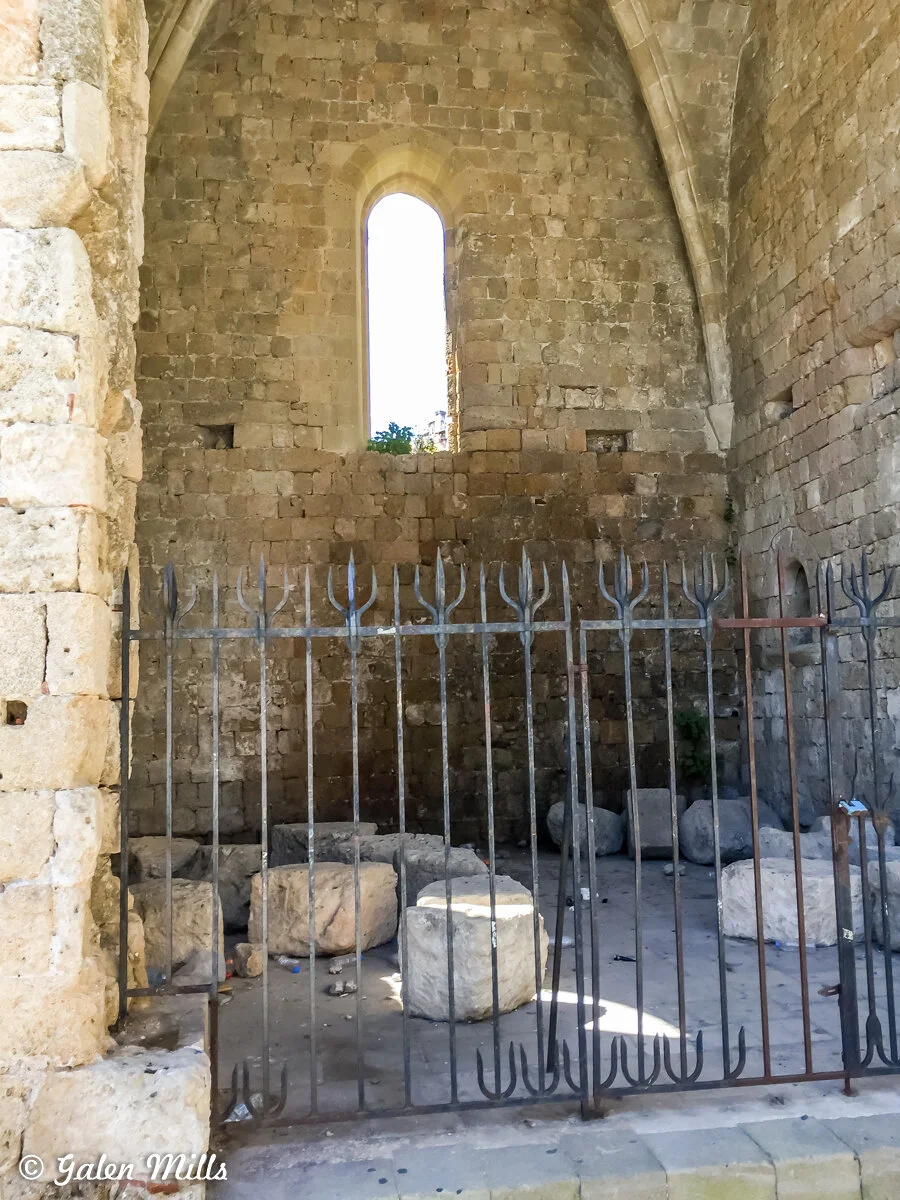 Interior of an old stone building with arched ceiling, narrow window, and iron gate. The floor has large stone blocks scattered around.