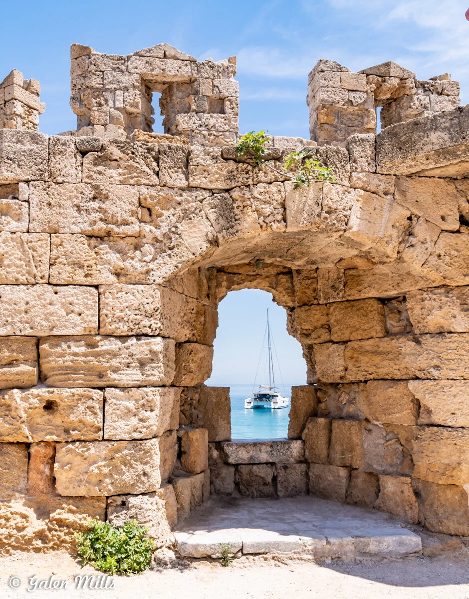 Ancient stone wall with arched opening, revealing a sailboat on a calm blue sea under a clear sky.